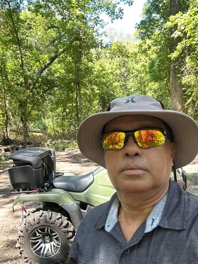 A rider in a sun hat taking a selfie beside his ATV on a wooded trail.