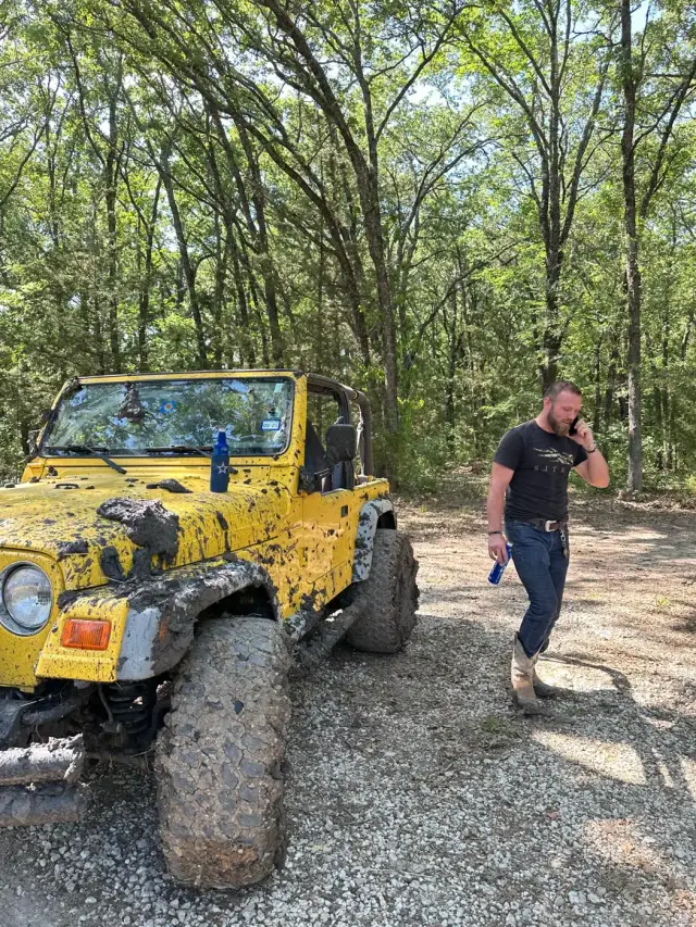 A muddy yellow Jeep parked on gravel with a rider on the phone beside it.