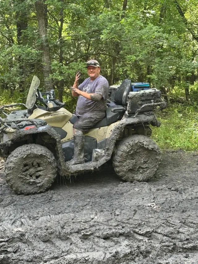 A rider covered in mud waving from his ATV in a muddy clearing.